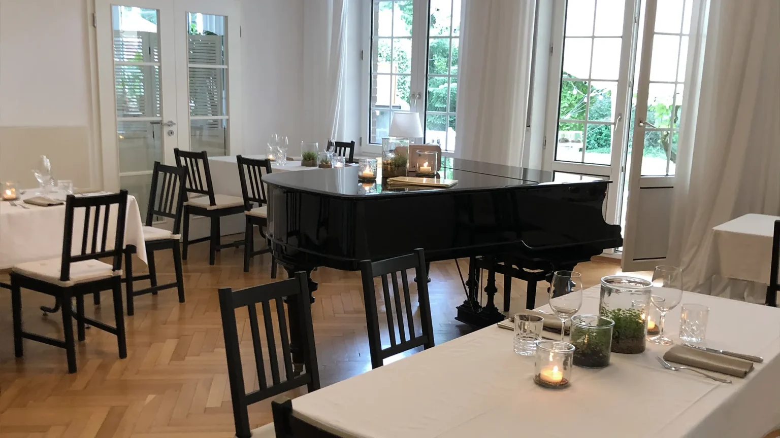 Einladend gedeckter Tische im Gastraum der Villa Schwenk mit weißem Tischtuch, schwarzen Stühlen und eleganter Dekoration. In der Mitte des Raumes ein schwarzer Flügel.