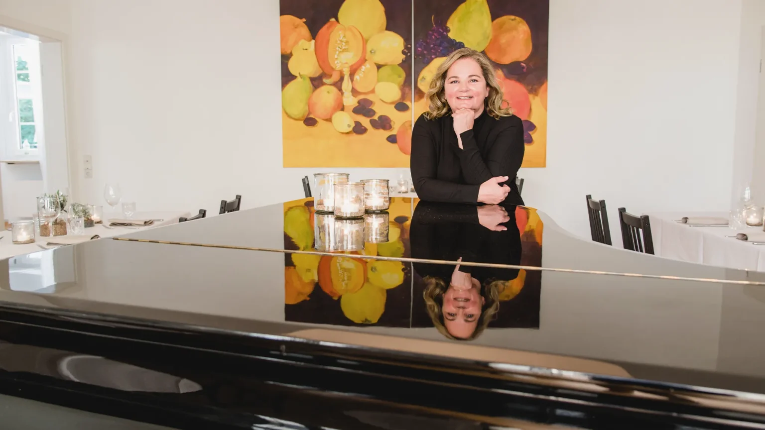 Gastgeberin Yvonne Schneider hinter einem schwarzen Flügel im Gastraum der Villa Schwenk. Im Hintergrund elegant gedeckte Tische.